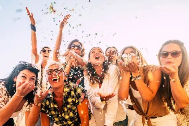 Photo of People having fun in party celebration friends concept - group of young and adult women all together laughing blowing coloured confetti - friendship and love for lifestyle with mixed active generations