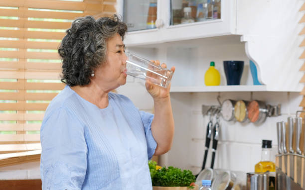 senior asian woman drinking water while standing by window in kitchen background, people and healthy lifestyles - thirsty imagens e fotografias de stock