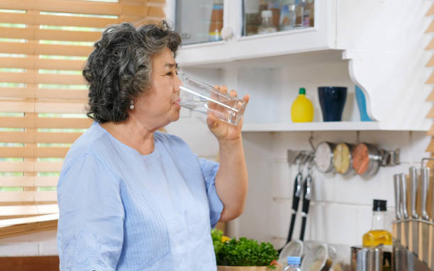 старшая азиатская женщина питьевой воды, стоя у окна на кухне фоне, люди и здоровый образ жизни - thirsty стоковые фото и изображения