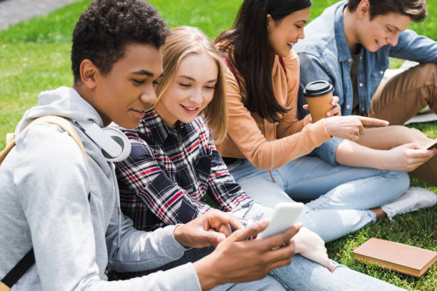 adolescenti sorridenti e felici seduti sull'erba e guardando lo smartphone - solo adolescenti foto e immagini stock