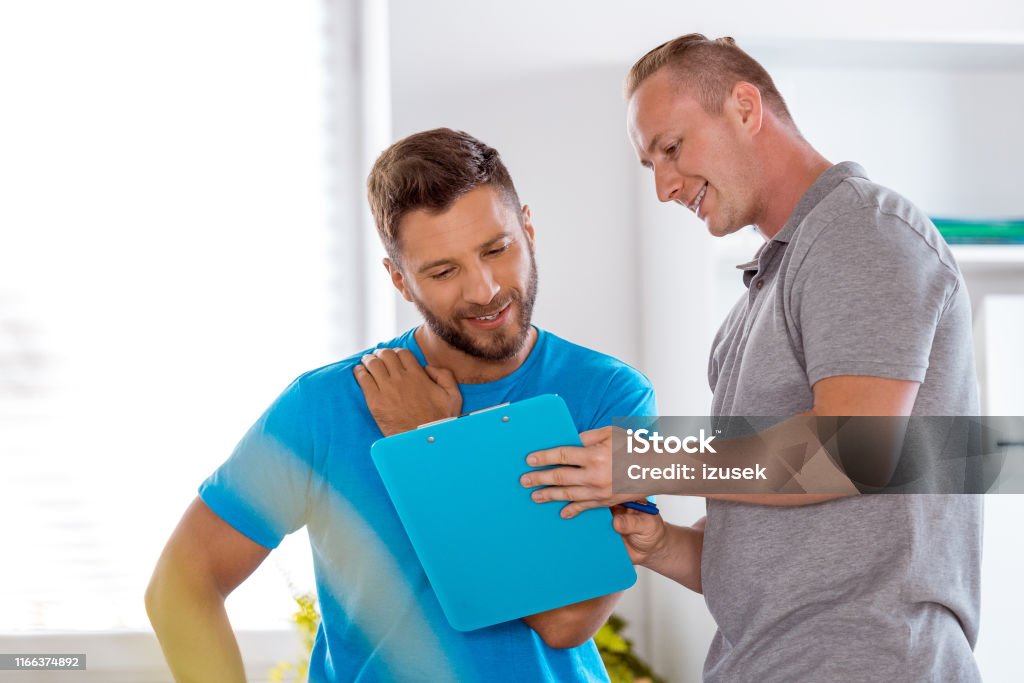 Therapist talking with a athlete in rehabilitation center Physical therapist talking with young male athlete in rehabilitation center and showing him his medical result on clipboard. Discussion Stock Photo