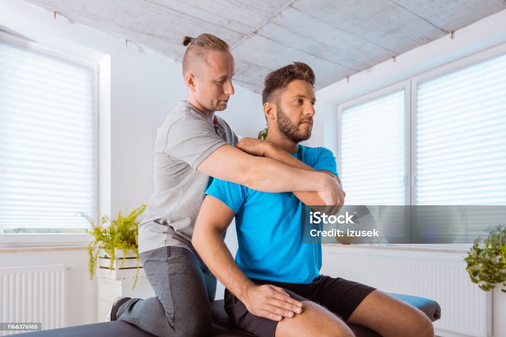 Therapist massaging young man at medical center Male physiotherapist giving massage to a young man at rehab center. Therapist massaging young man at medical center. Physical Injury Stock Photo