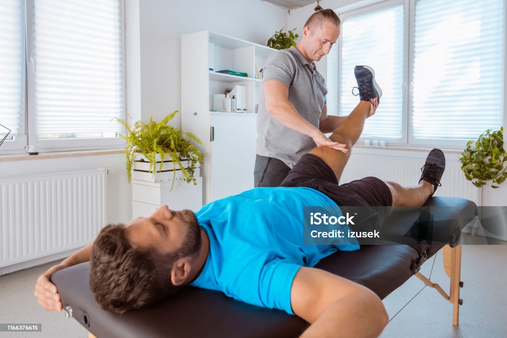 Therapist massaging young man at medical center Male physiotherapist giving massage to a young man at rehab center. Therapist massaging young man at medical center. Adult Stock Photo