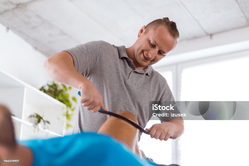 Therapist giving knee massage using a tool Therapist giving knee massage to young man using a tool at rehabilitation center Knee Stock Photo