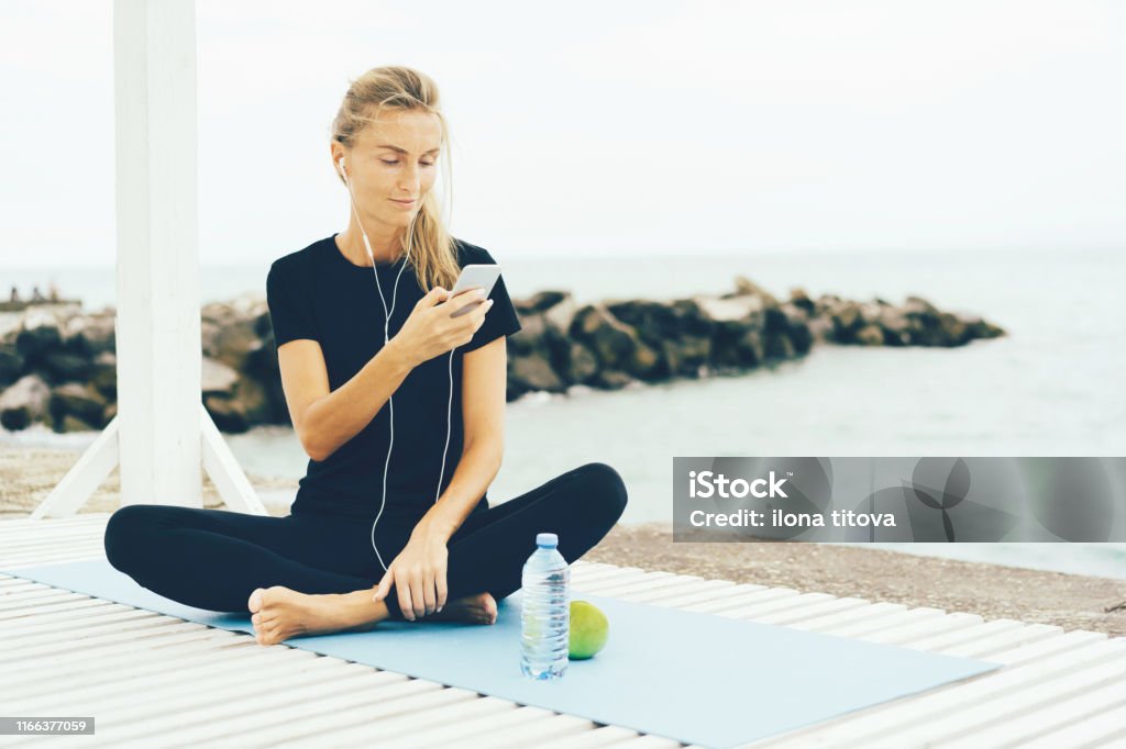Woman after workout with a phone in her hands Young sporty woman listening to music on smartphone after training outside on the seashore. Break after hard workout. Mail Stock Photo