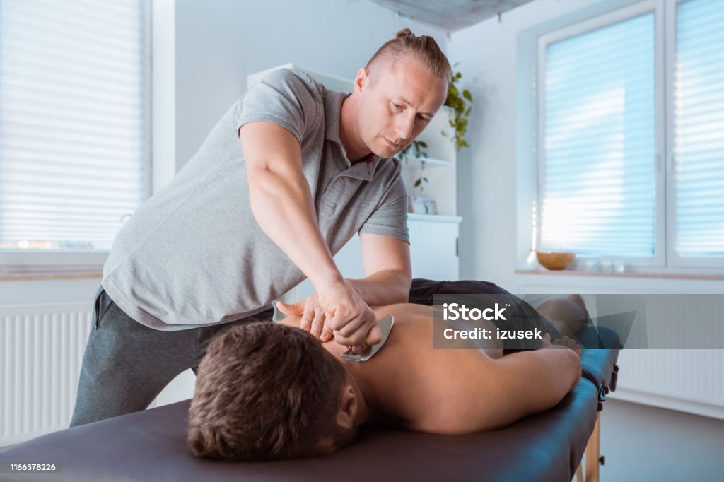 Professional therapist giving back massage with a tool Professional physical therapist giving back massage to a man using a tool at medical center Adult Stock Photo