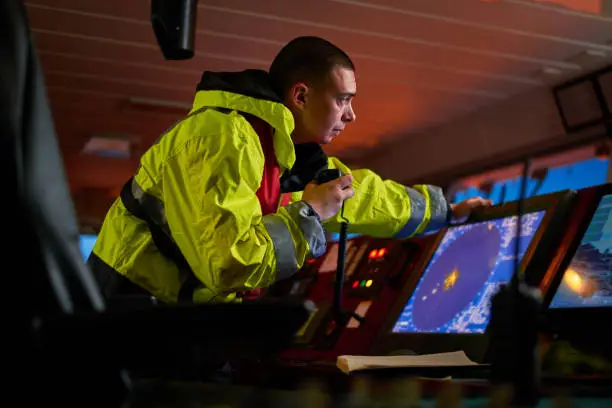 Navigator. pilot, captain as part of ship crew performing daily duties with VHF radio, binoculars on board of modern ship with high quality navigation equipment on the bridge on sunrise.