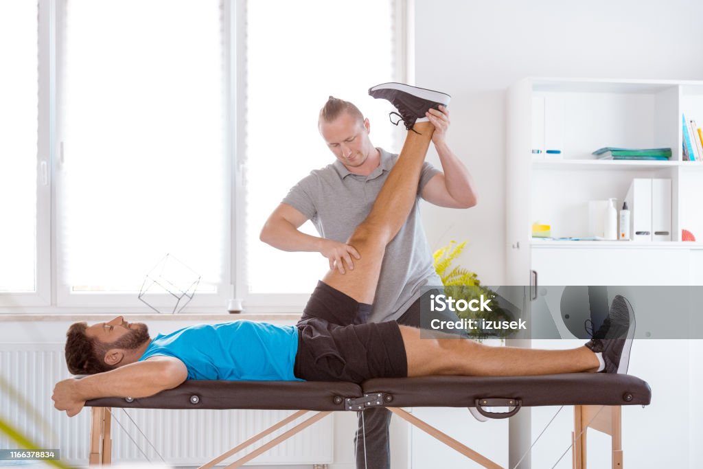 Physiotherapist assisting a young patient in recovery Young man lying on bed with his therapist stretching and examining his legs at rehabilitation center Physical Therapy Stock Photo