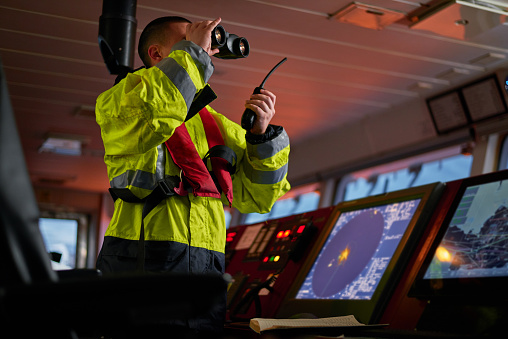 Navigator. pilot, captain as part of ship crew performing daily duties with VHF radio, binoculars on board of modern ship with high quality navigation equipment on the bridge on sunrise.