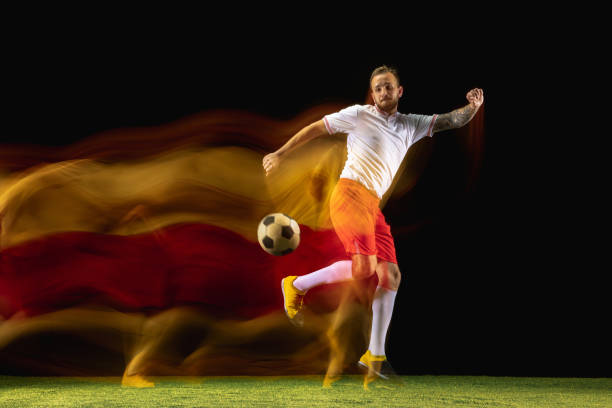 Burn. Young caucasian male football or soccer player in sportwear and boots kicking ball for the goal in mixed light on dark background. Concept of healthy lifestyle, professional sport, hobby.
