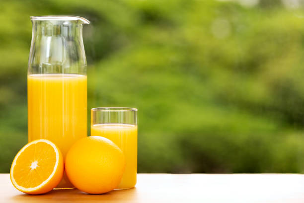 Orange juice in a jug and a glass with full and cut orange by a window opening to tree tops, shallow depth of field. large copy space. stock photo