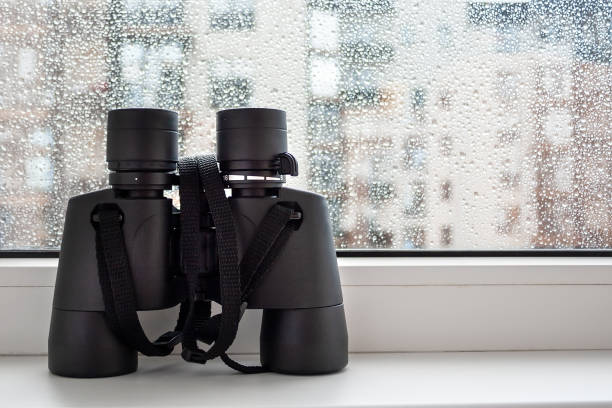 istock On a white windowsill by the window with raindrops on the glass is a black binoculars for observing neighbors. Fascinating observation of the environment from the window of the house 1166413451