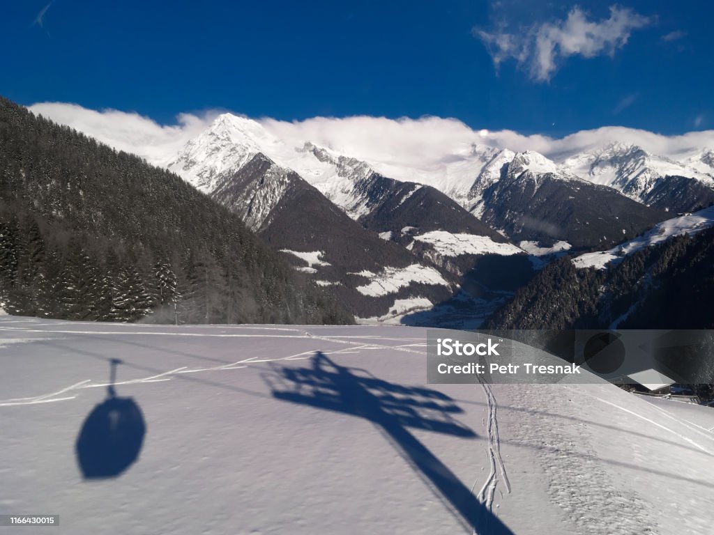 Cable car lift shadow silhouette on fresh snow with Aurina valley and mountains with foggy clouds on horizon below blue sky during sunny day in Speikboden ski resort in Italy Alto Adige - Italy Stock Photo