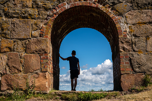 Summer view, silhouette of a young boy man leaning against an ancient old stone wall inside an arched gate looking at the blue sky. Varberg Fortress in Sweden. Horizontal composition.