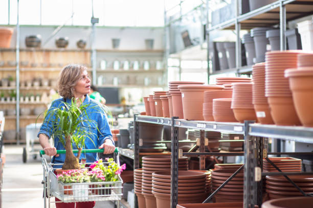 Mature Female Customer Looking at Garden Center Merchandise Woman in her 40s pushing shopping cart filled with flowering plants past shelves of terracotta pots in garden center. spanish flower pots stock pictures, royalty-free photos & images