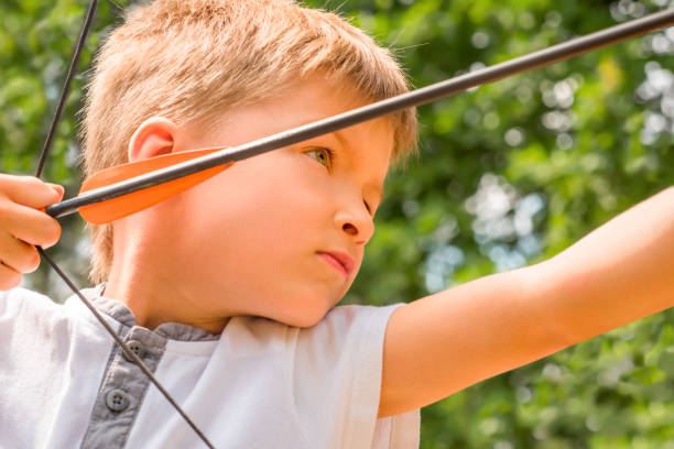 istock Boy with bow and arrow concentrated on target. Kid stared at target. Child directed arrow at a target. Bowman background. Children and sports. Physical training. Alternative schooling 1166443496