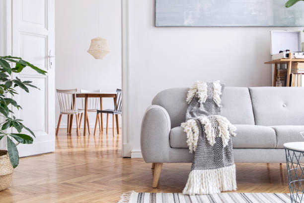 istock Modern scandianvian living room with design sofa with elegant blanket, coffee table and bookstand on the white wall. Brown wooden parquet. Concept of minimalistic interior. View on dining room. 1166445459