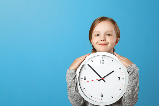 A cute little girl holding a big clock over a blue background. The concept of education, school, timing, time to learn. Copy space.