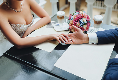 The groom holds the bride's hand in the cafe. Cups with cappuccino and bouquet from red rose. Wedding concept.
