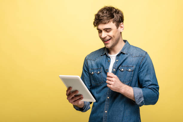 istock cheerful young man in denim shirt using digital tablet on yellow background 1166455331