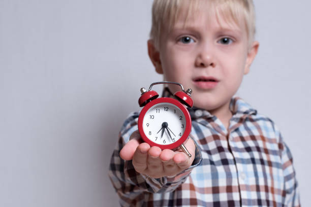 istock Little blond boy with a red alarm clock in his hands. Morning concept. Light background 1166458045