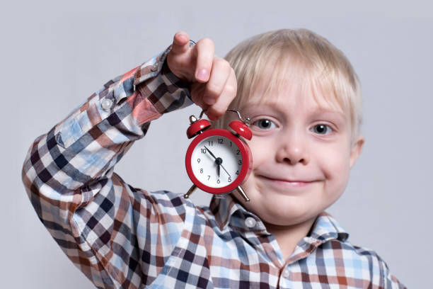 istock Little blond boy with a red alarm clock in his hands. Morning concept. Light background 1166458047