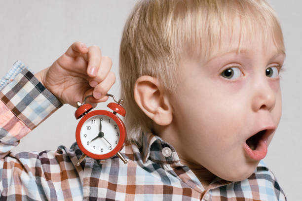 istock Little blond boy with a red alarm clock in his hands. Surprised face. Morning concept. Light background 1166458049