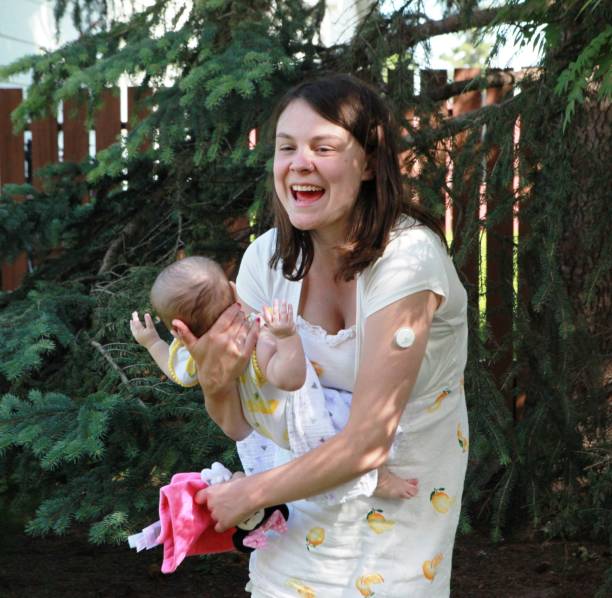 A medium over the shoulder shot of a cheerful young mother holding her baby girl while talking to a friend near a scenic lake. The woman is smiling brightly and dressed casually in a striped shirt and jeans, while the baby wears a pink outfit with matching shoes and harness. Captured in a candid moment, the scene reflects joy, friendship, and parenthood on a sunny day in the park.Videos are available similar to this scenario.