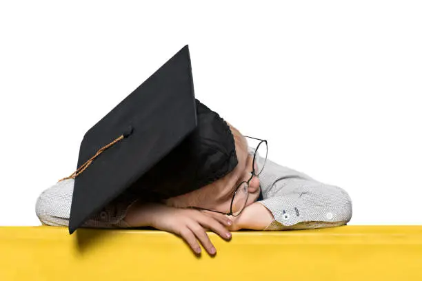 Photo of Blond boy in an academic hat and glasses asleep on the table. Isolate. School concept