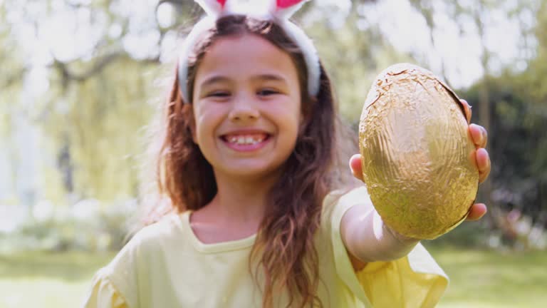 Portrait Of Girl Wearing Bunny Ears Holding Chocolate Egg On Easter Egg Hunt In Garden
