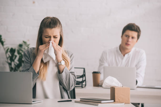 blonde businesswoman sneezing in tissue near coworker in office stock photo