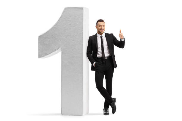 Young man in a black suit and tie leaning on number one and showing thumbs up Full length portrait of a young man in a black suit and tie leaning on number one and showing thumbs up isolated on white background leaning stock pictures, royalty-free photos & images