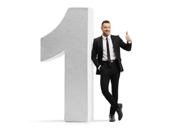 Full length portrait of a young man in a black suit and tie leaning on number one and showing thumbs up isolated on white background