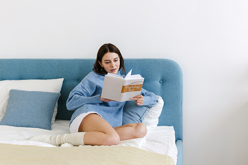Girl in a blue sweater reading a book on psychology sitting on the bed