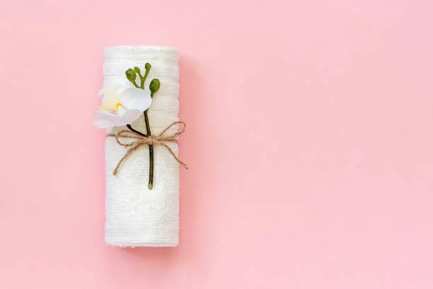 White towel roll tied with rope with sprig of orchid flower on pink paper background. Copy space Template for lettering text or your design.