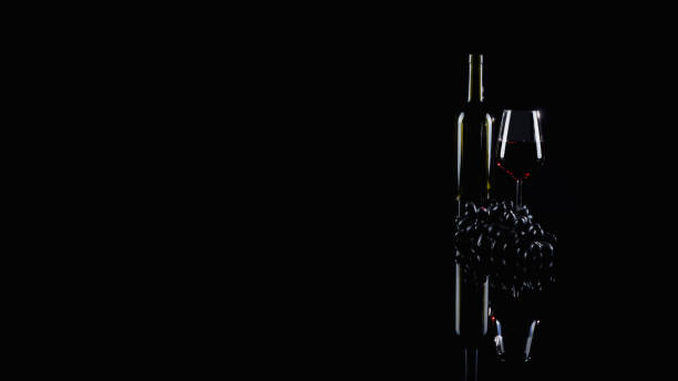 A glass and bottle of red wine and red grapes with reflaction on dark background