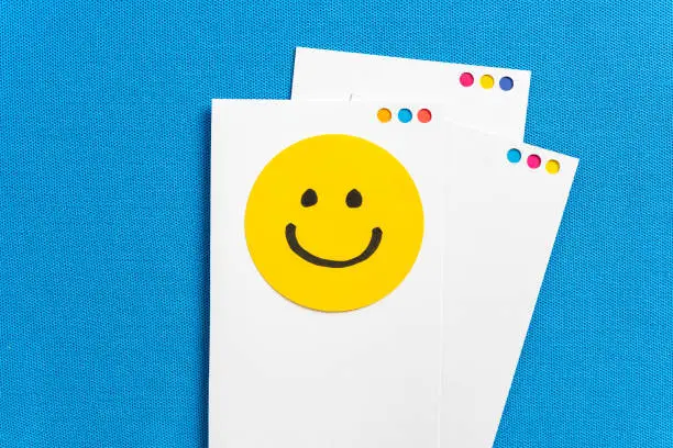 Photo of Concept of happy work, well-being, well done, feedback, employee recognition award. Paper white notes with yellow circle happy smiling face cartoon illustrated on blue texture background.