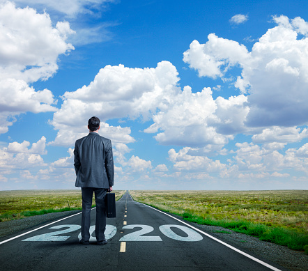 A businessman stands on and looks down a long, straight, rural road that has the year 2020 painted on it.  In the distance, as the road meets the horizon, puffy clouds punctuate the blue sky.