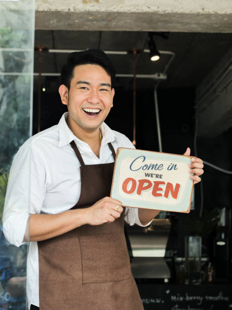 コーヒーショップの前でウェルカムボードを持つ陽気なアジアのバリスタの男。 - black sign holding vertical ストックフォトと画像
