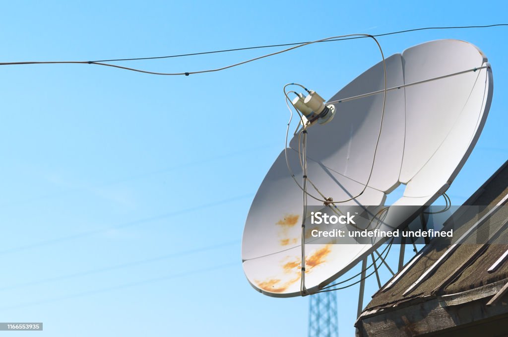 Old Satellite Television Antenna Dish on Top of a Barn Roof Covered with Waterproofing Tar Paper against Summer Blue Sky. Antenna - Aerial Stock Photo