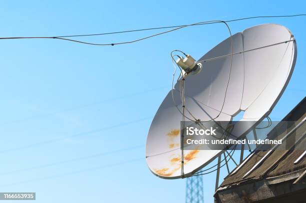 Old Satellite Television Antenna Dish On Top Of A Barn Roof Covered With Waterproofing Tar Paper Against Summer Blue Sky Stock Photo - Download Image Now
