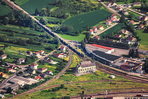 Aerial view on freight train arriving on Culoz town railroad track junction, with old building structure, in middle of French countryside in summer, Ain department, Auvergne-Rhone-Alpes region