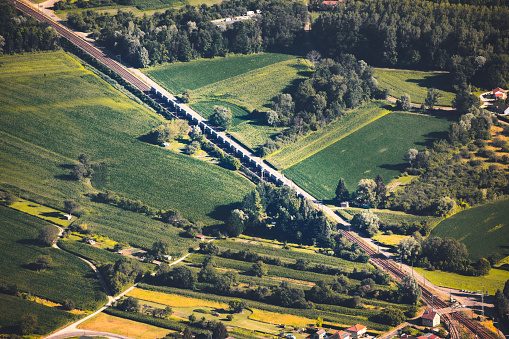 Horizontal color photography of aerial view on freight train crossing in diagonal the French countryside in summer, in middle of vibrant green fields. Shot from Grand Colombier mountain top during a sunny summer day, in Bugey mountains, in Ain department not far from Jura and Savoie border near Culoz city, Auvergne-Rhone-Alpes region in France (Europe).