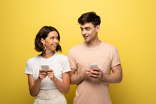 positive man and woman looking at each other while using smartphones on yellow background