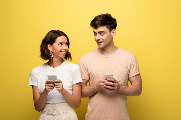 положительные мужчина и женщина смотрят друг на друга при использовании смартфонов на желтом фоне - лицом к лицу стоковые фото и изображения