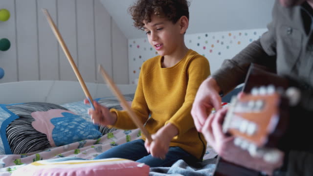 Single Father Playing Guitar With Son Who Drums On Cushion In Bedroom