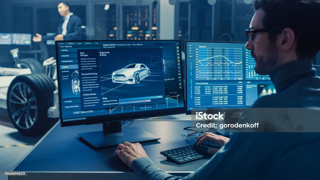 Professional Engineer Works on a Computer with a 3D CAD Software and Tests the Electric Car Chassis Prototype with Wheels, Batteries and Engine Standing in a High Tech Development Laboratory. Car Stock Photo