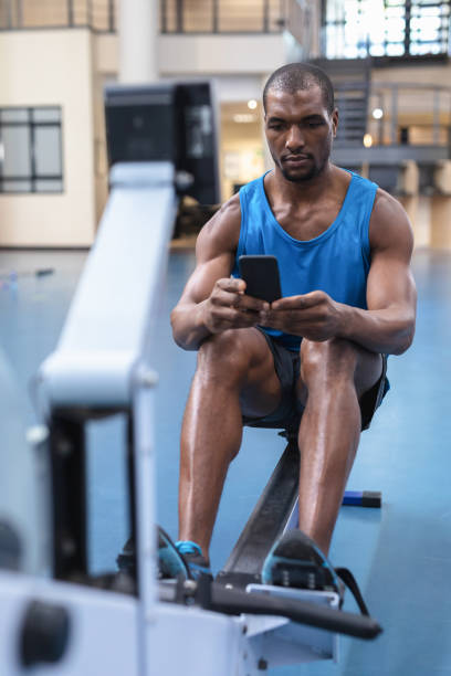 istock Man using mobile phone while exercising on rowing machine in fitness center 1166605866