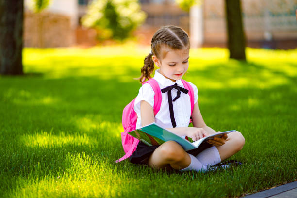 istock Little school girl with pink backpack sitting on grass after lessons and read book or study lessons, thinking ideas, education and learning concept 1166607345