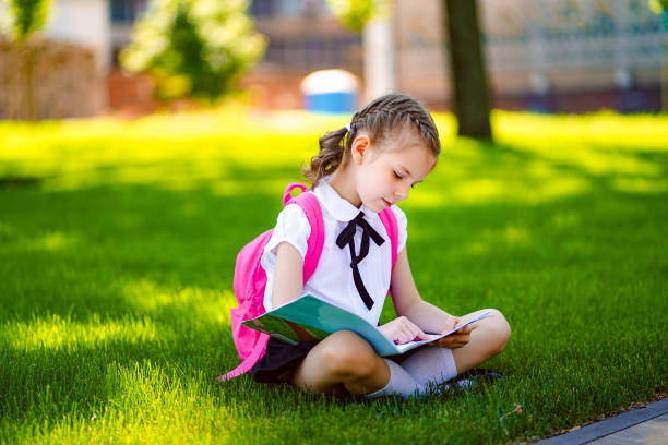 istock Little school girl with pink backpack sitting on grass after lessons and read book or study lessons, thinking ideas, education and learning concept 1166607370